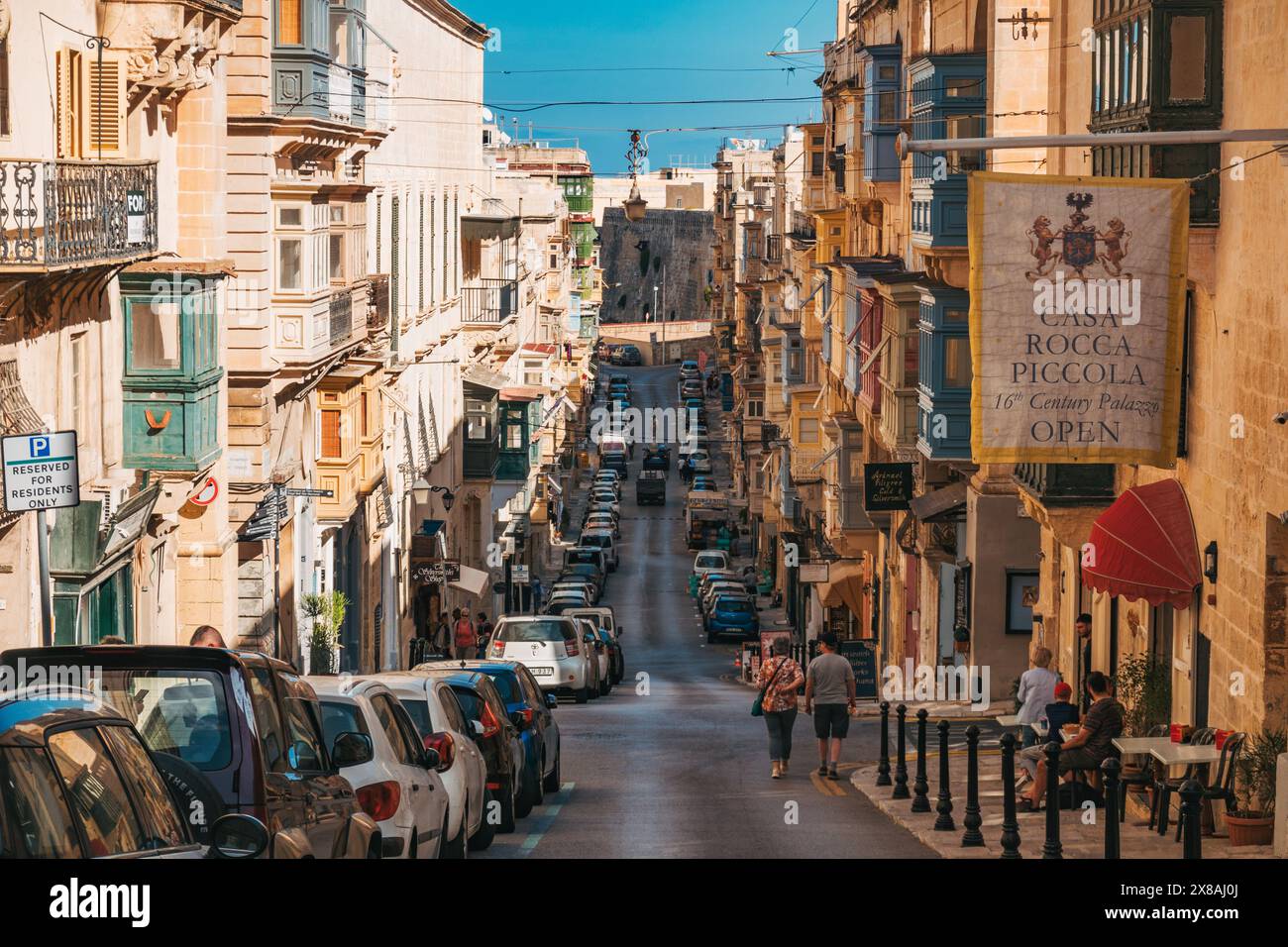 Una vivace strada a la Valletta presenta un'architettura affascinante e fiancheggiata da auto parcheggiate sotto un cielo azzurro Foto Stock