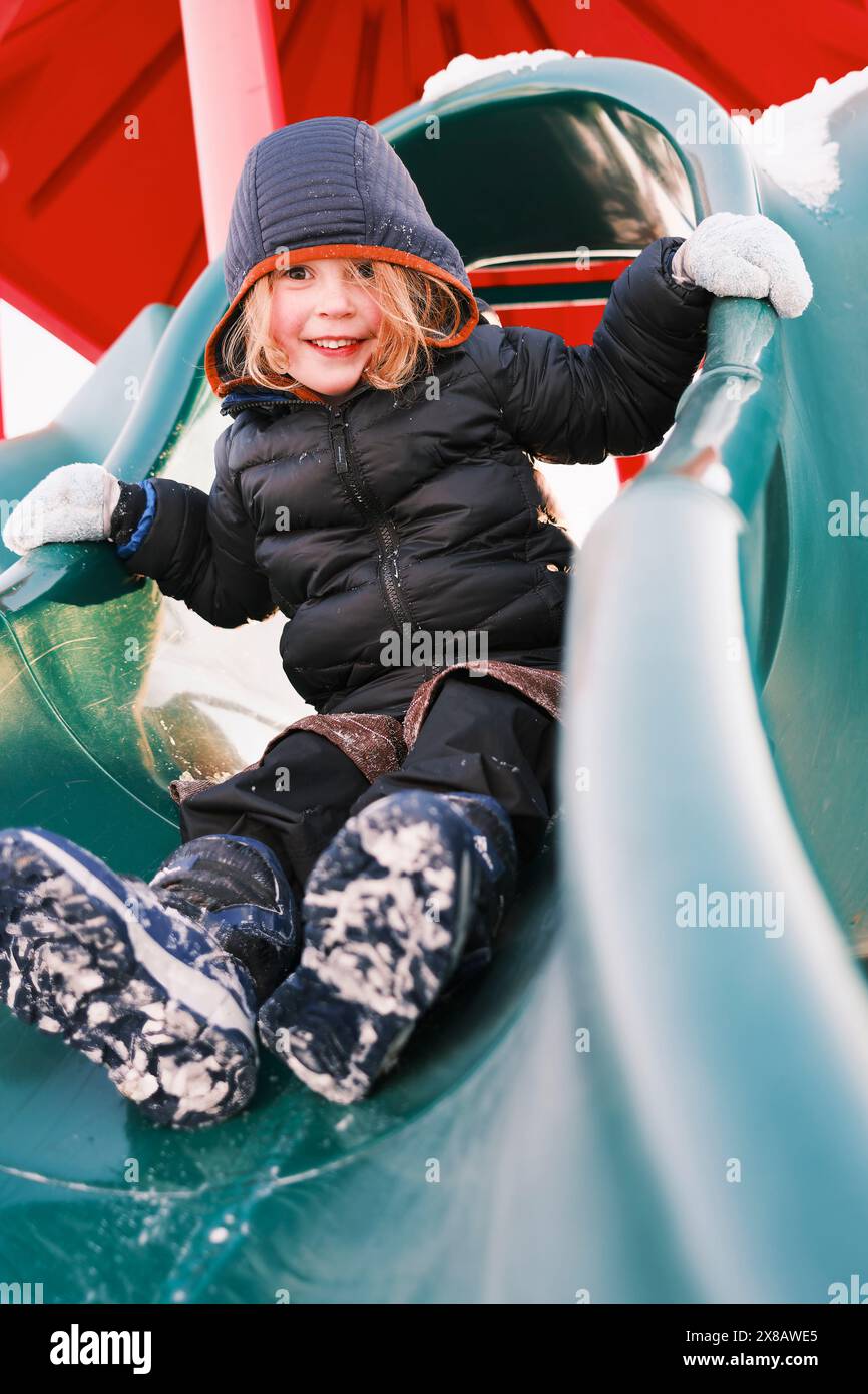 Bambino felice che gioca sullo scivolo nel parco giochi innevato Foto Stock