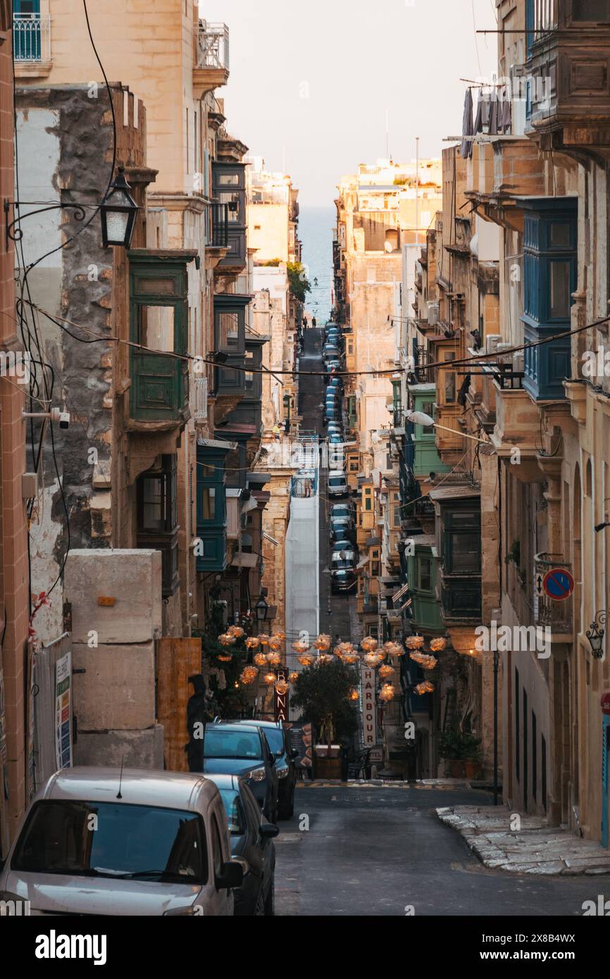 Una strada stretta e inclinata attraversa il terreno collinare di la Valletta, Malta, fiancheggiata da edifici storici e caratterizzata da auto parcheggiate e luci a corde Foto Stock