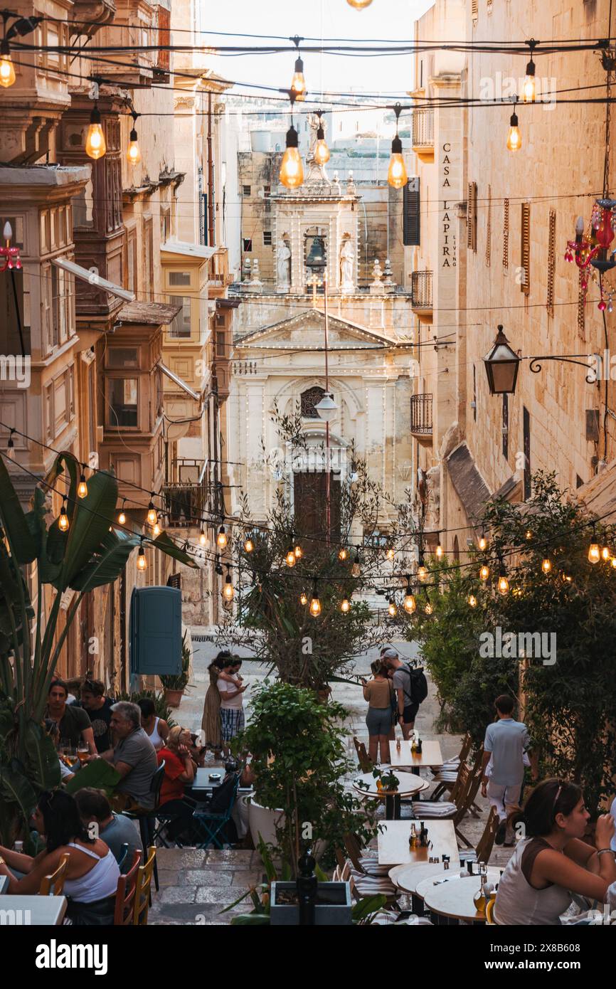 Le persone cenano all'aperto ai tavoli in una stradina stretta di la Valletta, Malta, illuminata da luci a corda Foto Stock