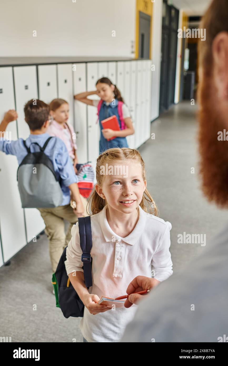 i bambini controllano con ansia gli armadietti in un corridoio vivace, guidato da un insegnante maschile in un luminoso ambiente scolastico. Foto Stock