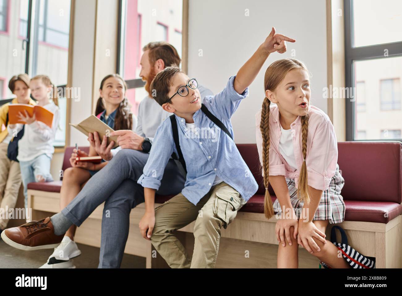 Un gruppo di bambini siede con attenzione su una panchina mentre un insegnante maschile li istruisce in un ambiente luminoso e vivace in classe. Foto Stock