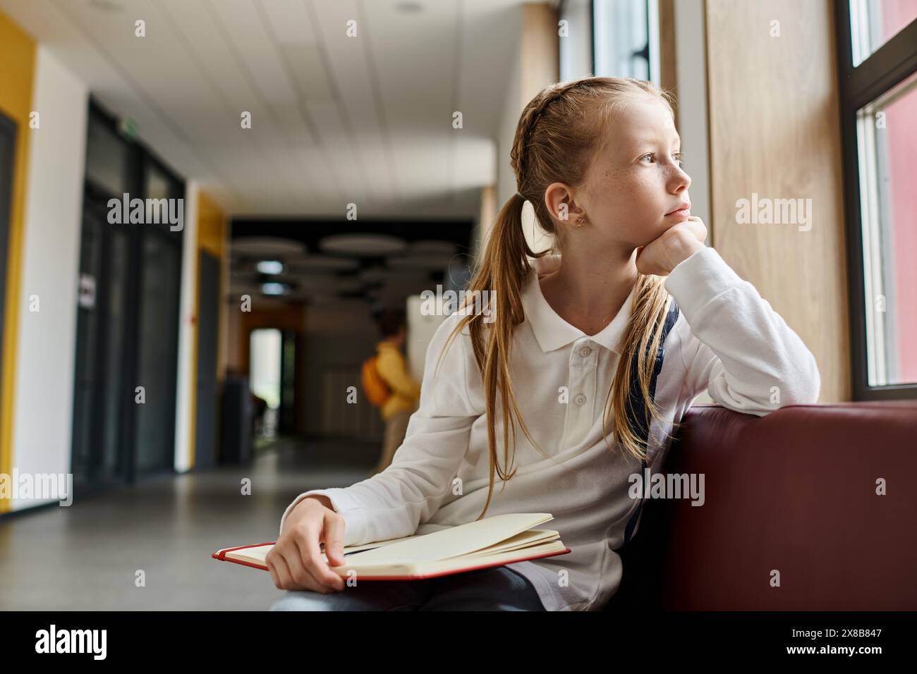 Una giovane ragazza con lunghi capelli scuri siede da sola su una panchina di legno, immersa in un libro, profondamente nel pensiero. Foto Stock