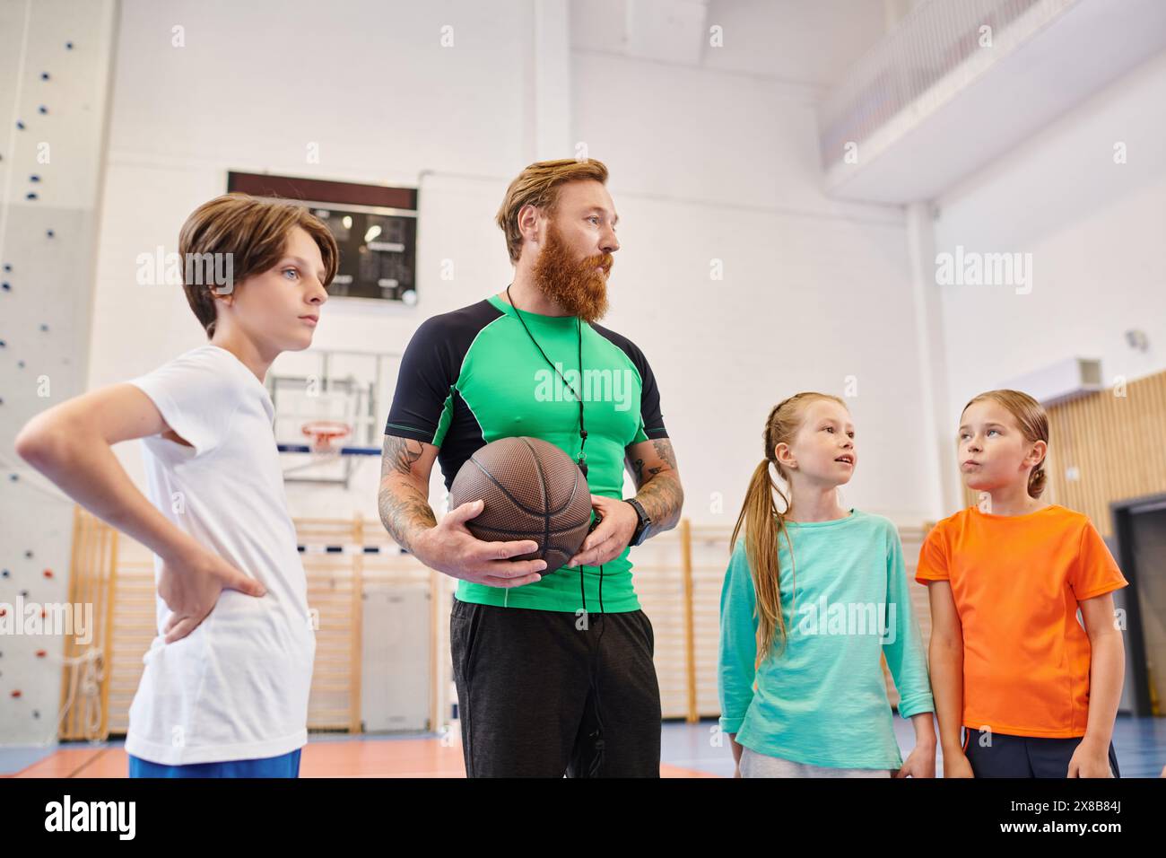 Un uomo che tiene un pallacanestro davanti a un gruppo di bambini diversi ed entusiasti in un ambiente luminoso in classe, che insegna e ispira loro. Foto Stock