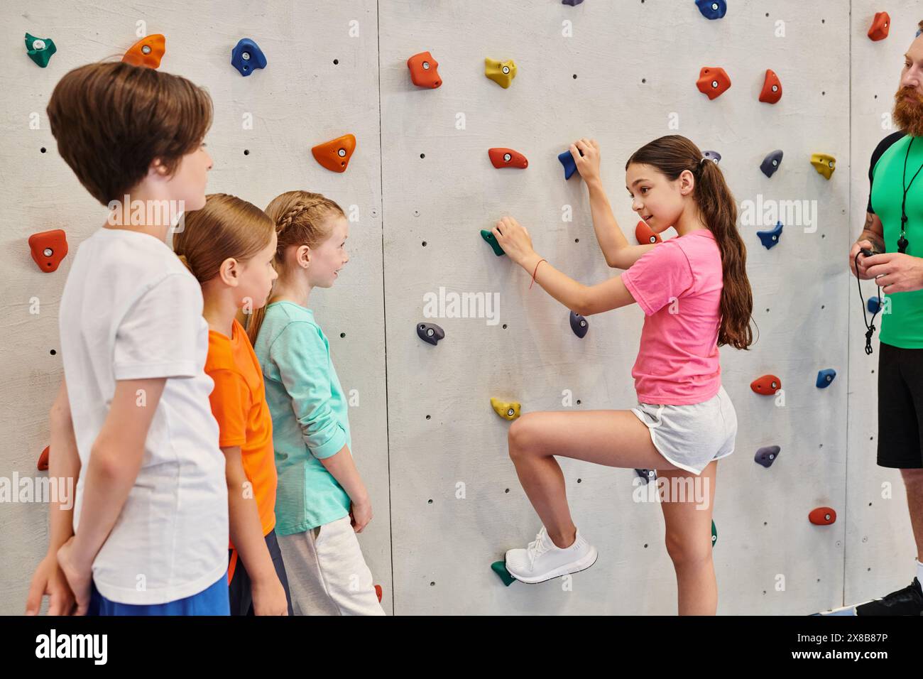 Un uomo insegna a un gruppo eterogeneo di bambini come arrampicarsi su un muro di roccia in un ambiente vivace ed energico. Foto Stock