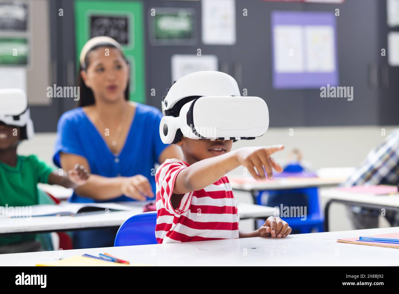 A scuola, in classe, un'insegnante birazziale femminile osserva gli studenti che indossano occhiali VR Foto Stock