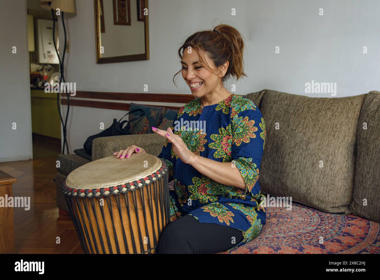 Una donna latina adulta e felice seduta a casa che esprime se stessa attraverso la musica africana con un tamburo djembe che suona musica africana a percussioni. Lei è sorridente Foto Stock