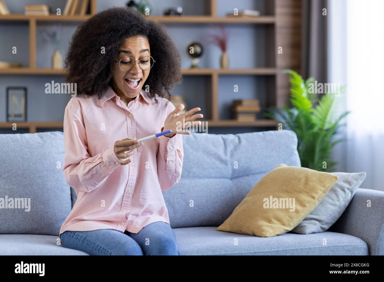 Una donna in salotto reagisce felicemente a un test di gravidanza positivo, seduto comodamente su un divano, esprimendo gioia con un sorriso. Gli interni moderni aggiungono un'atmosfera intima Foto Stock