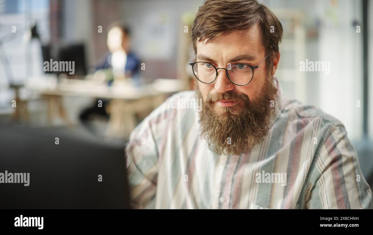 Ritratto di giovane uomo caucasico barbucato con gli occhiali concentrati e che lavora al computer in un ufficio luminoso e spazioso. Amministratore maschio sorridente dopo aver completato con successo un'attività. Foto Stock