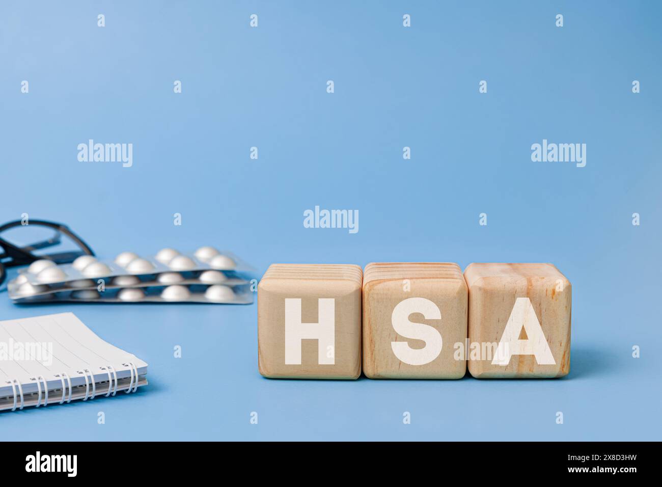 Blocco di legno con testo HSA che significa salute conto di risparmio con occhiali, notebook, pillole dietro su sfondo blu Foto Stock