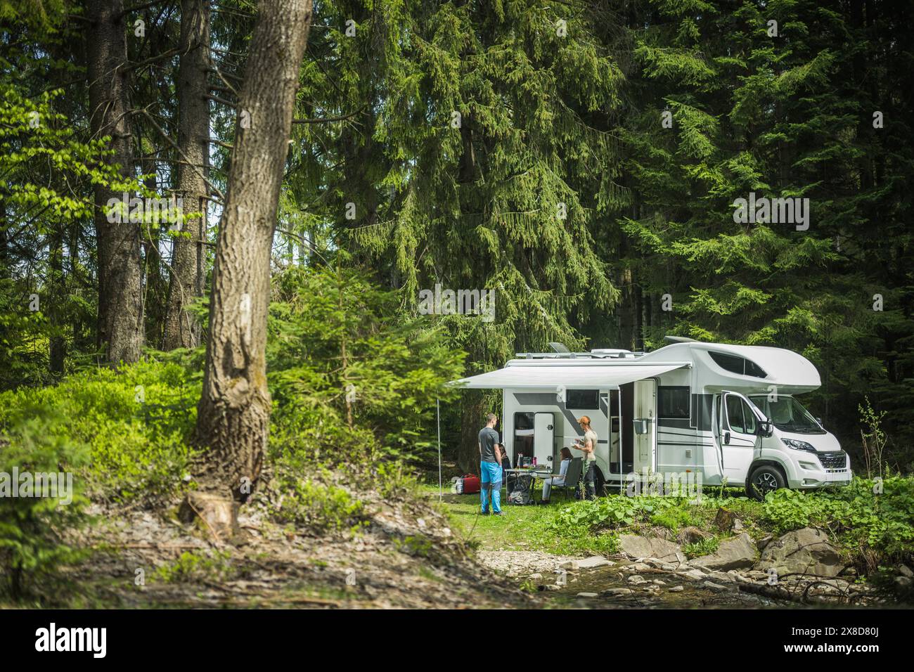 Amici in piedi di fronte a un camper parcheggiato, che si guardano l'un l'altro. Il camper è parcheggiato da un tranquillo lago, con alberi sullo sfondo. Foto Stock