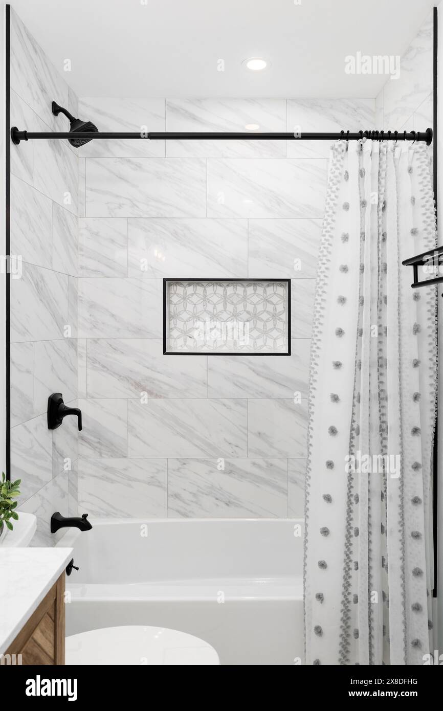 Una bellissima doccia con pareti piastrellate in marmo, un ripiano di nicchia in piastrelle a mosaico, soffione doccia e rubinetto neri e un mobile da toilette in legno. Foto Stock