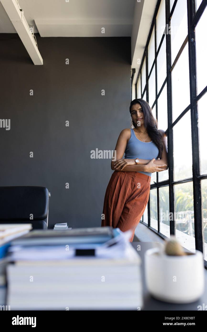 Nel moderno ufficio, donna indiana in piedi davanti a una grande finestra, appoggiata alla cornice, copia spazio. Ha lunghi capelli neri, pelle media, indossa un serbatoio blu Foto Stock