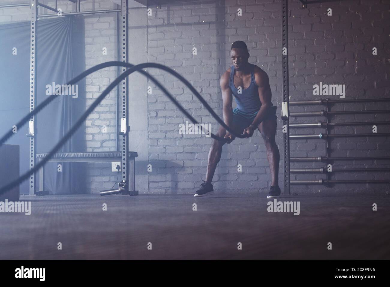Uomo afroamericano forte e in forma con corde da battaglia in palestra. Palestra in stile industriale con scaffali in metallo e pareti in mattoni che creano un robusto backgroun Foto Stock