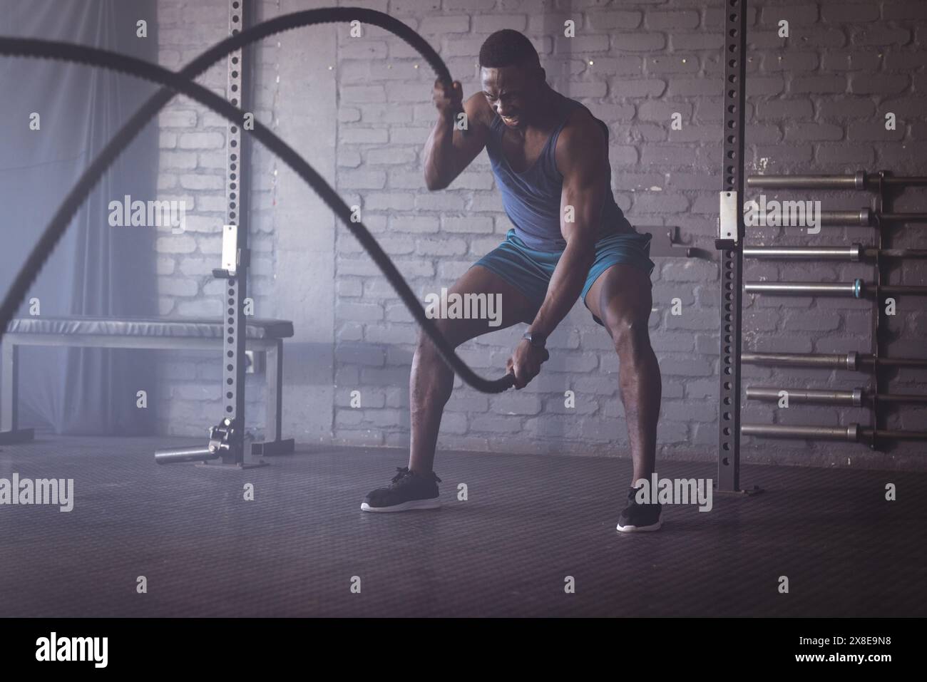 Uomo afroamericano forte e in forma con corde da battaglia in palestra. E' circondato da attrezzature da palestra e pareti in mattoni, creando un intenso bancomat di allenamento Foto Stock