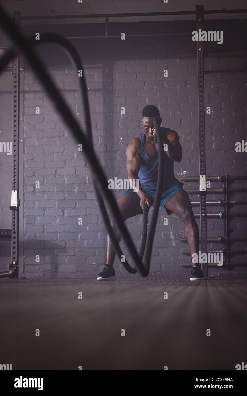 Uomo afroamericano forte e in forma in palestra con corde da battaglia. Si trova in una palestra moderna con pareti in mattoni e attrezzature per l'allenamento in background, concentrazione Foto Stock