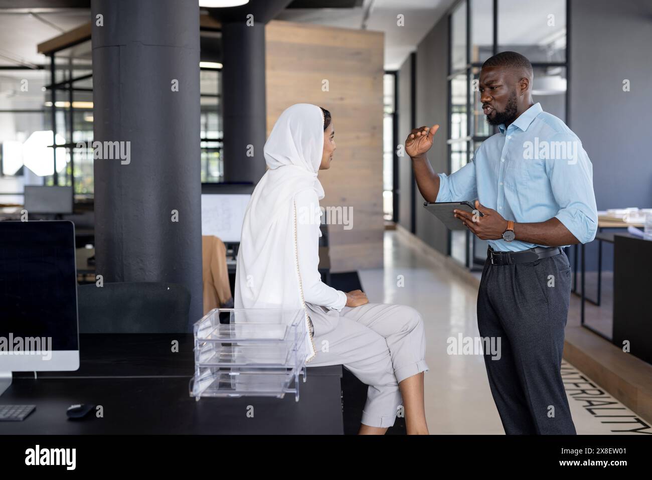 In un ufficio moderno, un team eterogeneo con colleghi afroamericani e indiani che parlano. Uomo afroamericano con capelli corti e barba che tiene la tavoletta, in Foto Stock