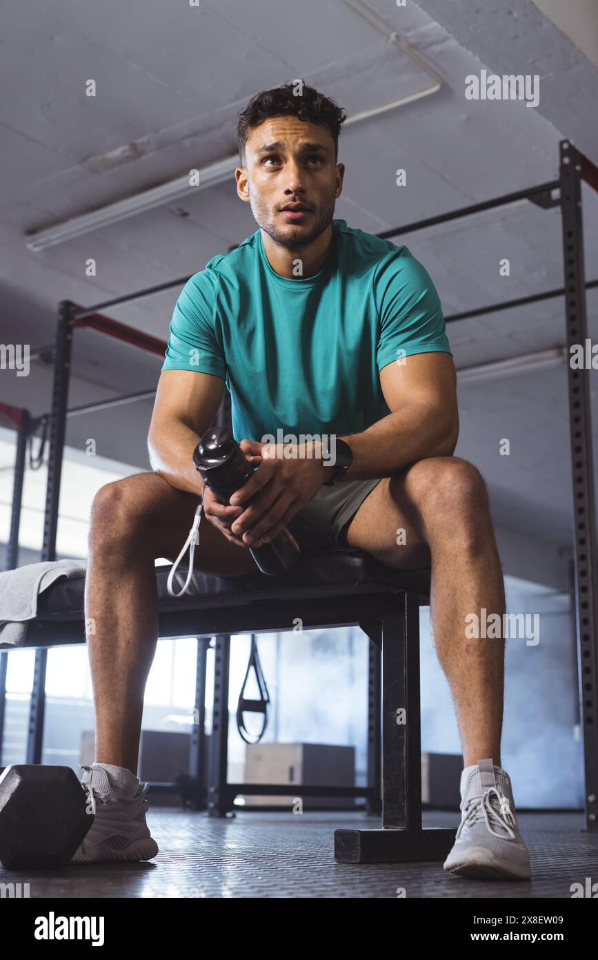 In palestra, uomo birazziale seduto sulla panchina della palestra tenendo una bottiglia d'acqua, sembra concentrato. Attrezzature da palestra e pesi sullo sfondo, creando un allenamento dedicato a. Foto Stock