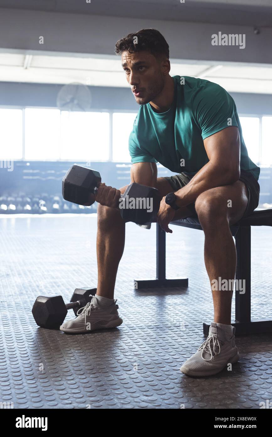 Uomo birazziale forte e in forma in palestra che solleva manubri seduto sulla panchina Foto Stock Uomo birazziale forte e in forma in palestra che solleva manubri seduto sulla panchina Foto Stock