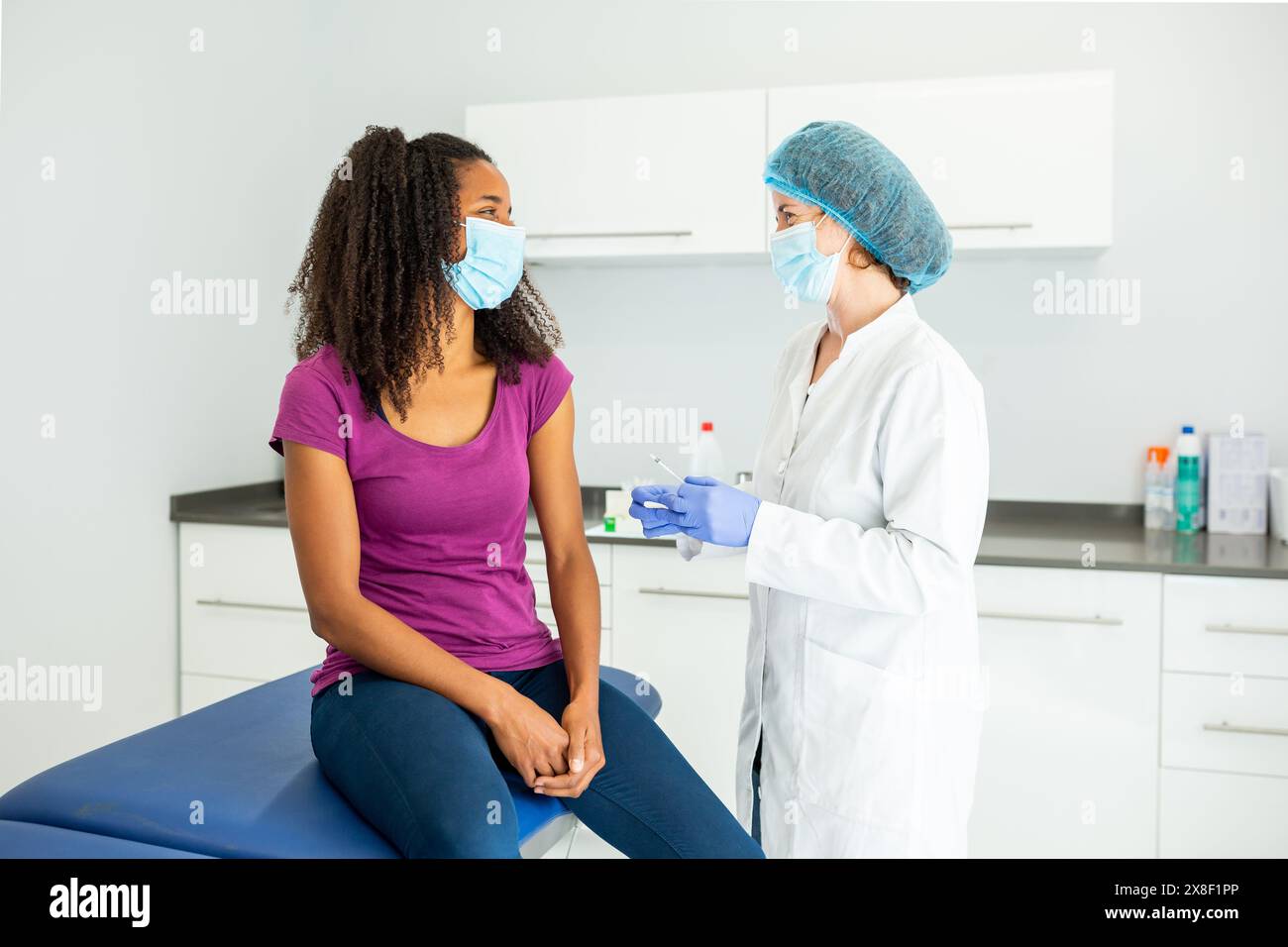 Medico che si sta preparando a somministrare un vaccino ad un paziente seduto su un tavolo d'esame. Entrambi gli individui indossano maschere e sono impegnati in un convers Foto Stock