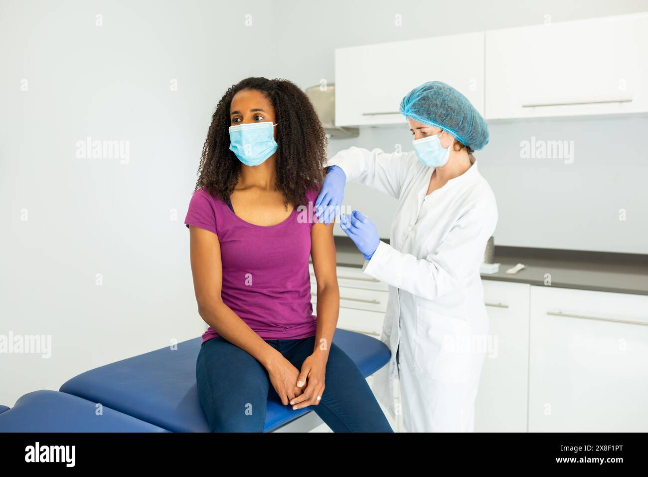 Medico che si sta preparando a somministrare un vaccino ad un paziente seduto su un tavolo d'esame. Entrambi gli individui indossano maschere e sono impegnati in un convers Foto Stock