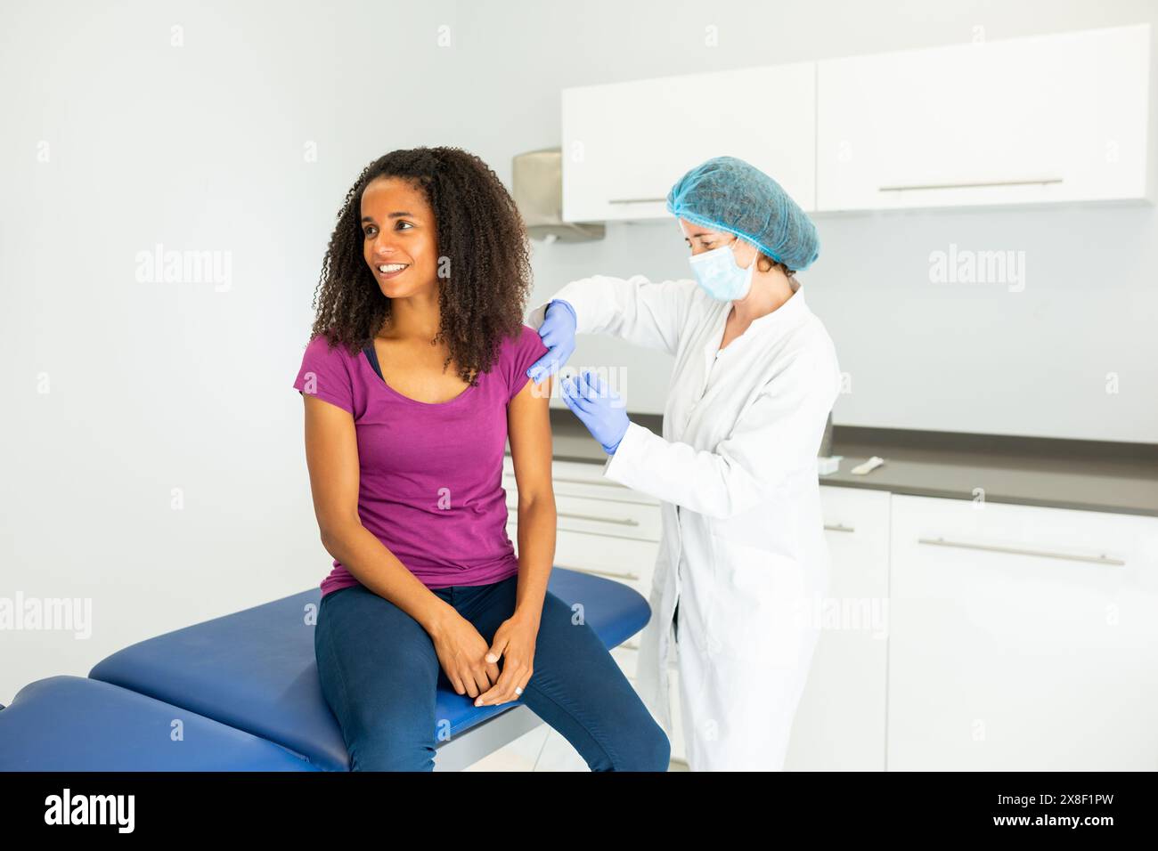 Medico che si sta preparando a somministrare un vaccino ad un paziente seduto su un tavolo d'esame. Entrambi gli individui indossano maschere e sono impegnati in un convers Foto Stock