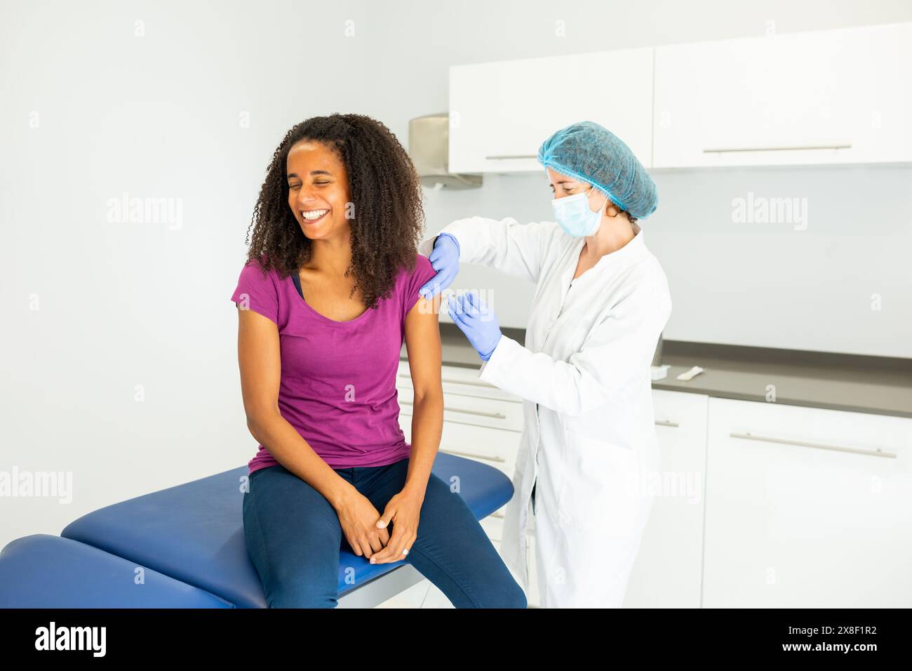 Medico che si sta preparando a somministrare un vaccino ad un paziente seduto su un tavolo d'esame. Entrambi gli individui indossano maschere e sono impegnati in un convers Foto Stock