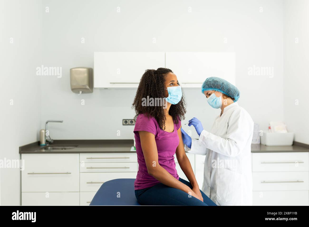 Medico che si sta preparando a somministrare un vaccino ad un paziente seduto su un tavolo d'esame. Entrambi gli individui indossano maschere e sono impegnati in un convers Foto Stock
