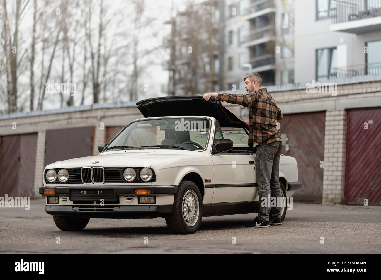 Riga, Lettonia - 1 aprile 2024: Un uomo che piega il tetto della classica BMW E30 cabriolet bianca. Vecchi garage e moderno edificio sullo sfondo. Foto Stock