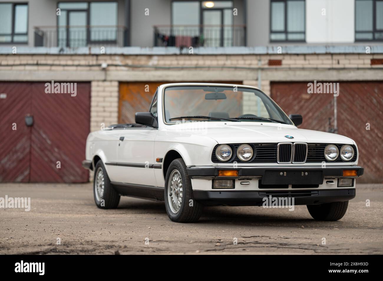 Riga, Lettonia - 1 aprile 2024: La classica BMW E30 con capote bianca con tetto aperto. Un vecchio garage e un edificio moderno sullo sfondo. Foto Stock