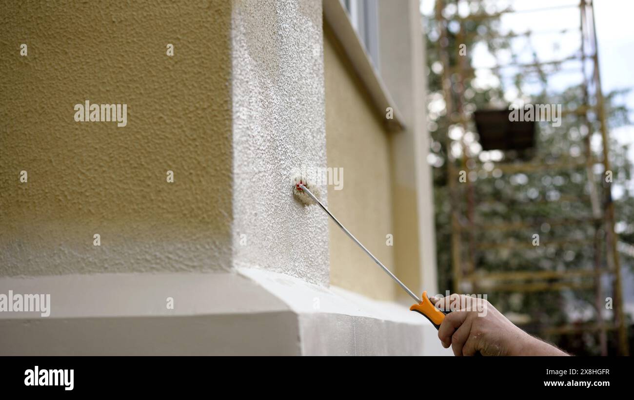 Dipingendo la facciata di una casa con un rullo con manico lungo. Lavoratore che tiene in mano il pennello, rullo per pittura giallo sulle pareti della casa, artigianato e concetto di ristrutturazione Foto Stock