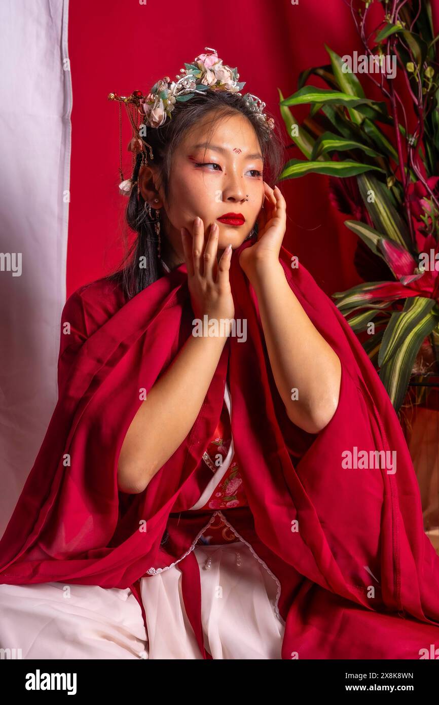 Una donna con la veste rossa si siede sul pavimento con le mani sul viso. Indossa un cerchietto con fiori e il rossetto rosso. L'immagine ha un caldo Foto Stock