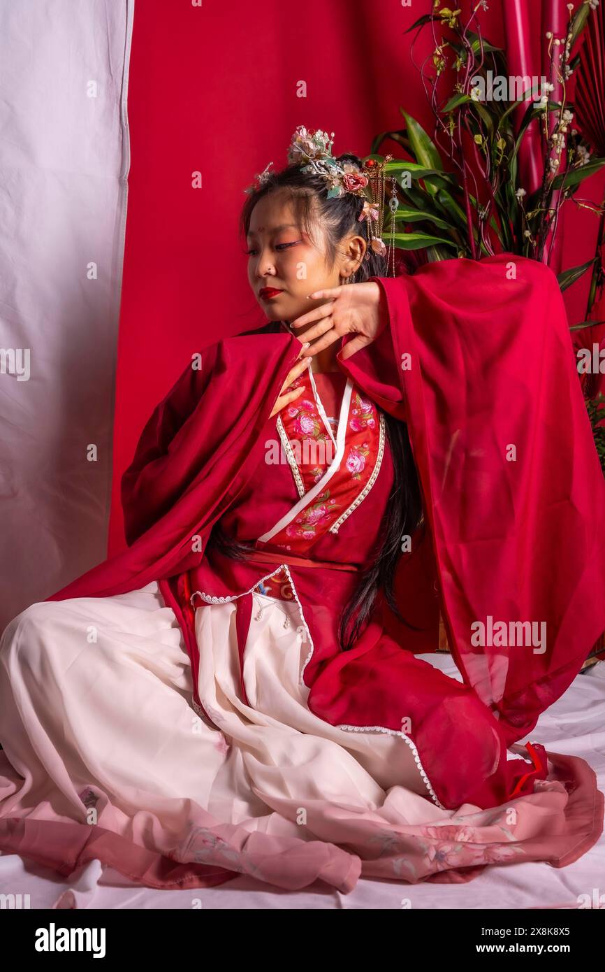Una donna con un vestito rosso e bianco è seduta su una superficie bianca. Indossa un abito tradizionale cinese e ha le mani sui fianchi. L'immagine Foto Stock