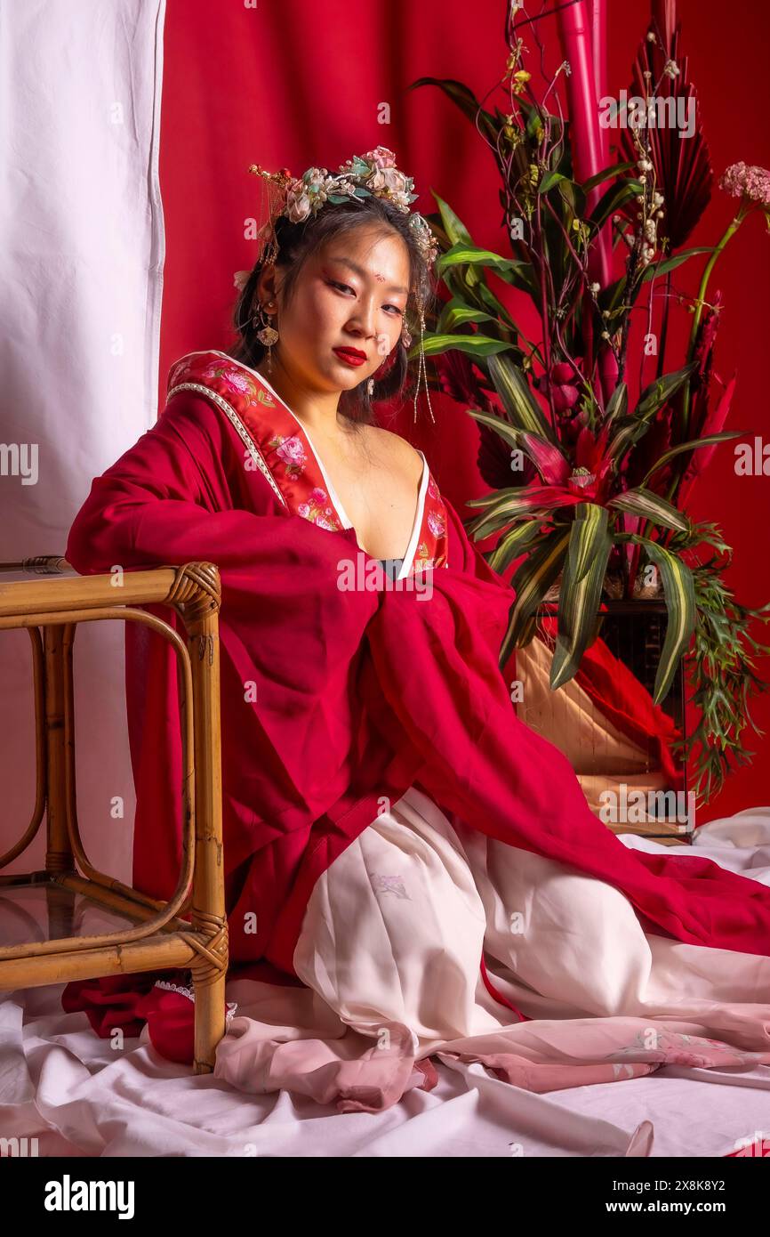 Una donna con un abito rosso e bianco siede su una sedia di legno. La donna è seduta di fronte a una pianta in vaso Foto Stock