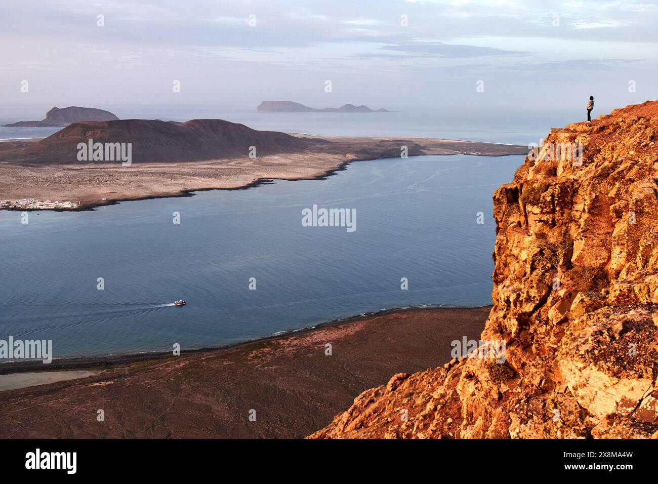 Una figura solitaria si erge su un'aspra scogliera, guardando il mare turchese e la natura selvaggia incontaminata che si estende lungo la costa lontana. Foto Stock