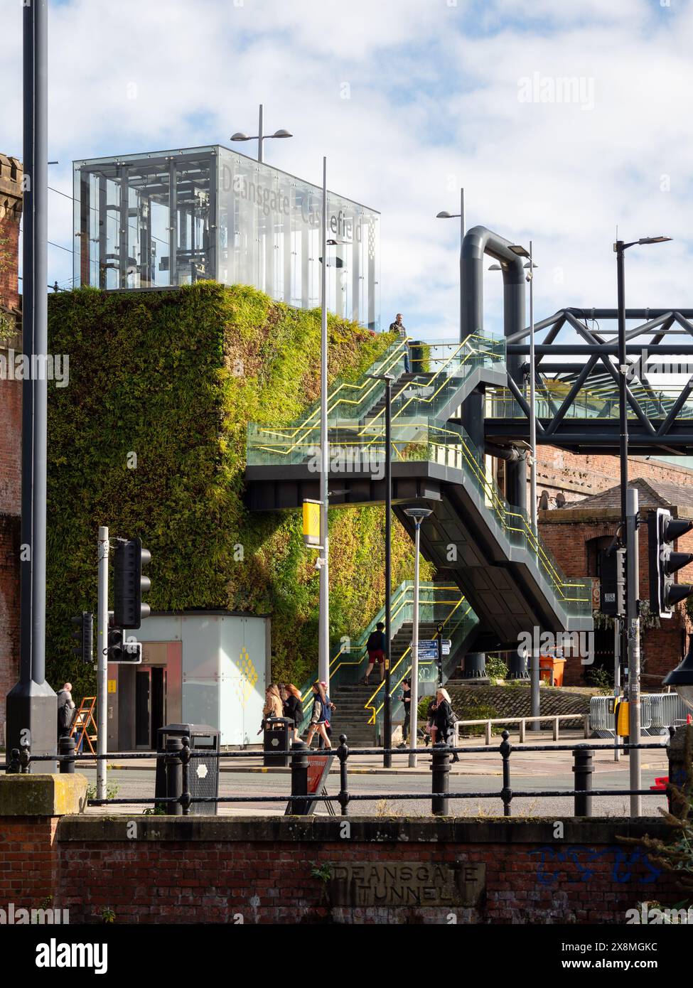 Manchester Regno Unito 26 maggio 2024. Ambiente urbano moderno con giardino verticale, ponte pedonale, ascensore in vetro e persone che camminano sotto il cielo azzurro Foto Stock