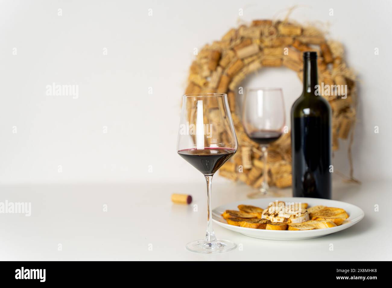 Bicchieri di vino rosso, bottiglia di vino e formaggio Brie al forno con pane tostato su un piatto bianco su un tavolo bianco con sfondo bianco e una corona di vino decorato Foto Stock