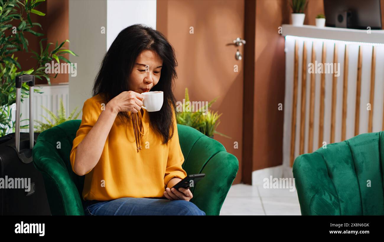Donna viaggiatrice in attesa di registrarsi in un hotel a cinque stelle, che utilizza lo smartphone per controllare le app dei social media e passare il tempo. Ospiti asiatici che arrivano presto al rifugio di vacanza, rilassandosi dopo un lungo volo. Foto Stock