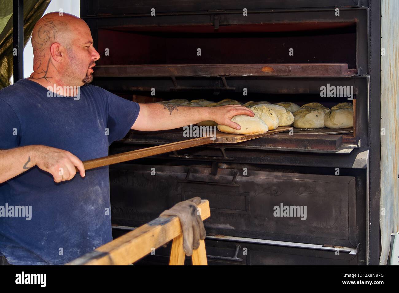 Viladecans, Barcellona, Spagna - 26 maggio 2024: Un fornaio tatuato gestisce un forno industriale per cuocere pane artigianale, mostrando abilità culinarie e tradizione. Foto Stock