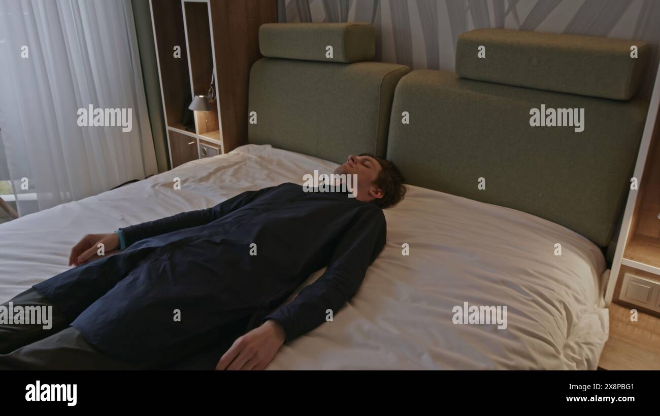 Giovane uomo sdraiato vestito con abiti su un letto incosciente o che dorme esausto. Fermo di scorta. Uomo caucasico sdraiato sul letto in una stanza con gli occhi chiusi. Foto Stock