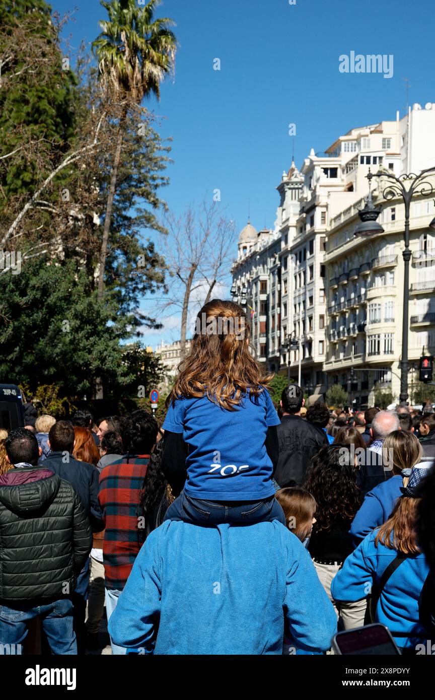 Una giovane ragazza siede sulle spalle di un adulto in mezzo alla folla durante il festival las fallas, con edifici storici e palme sullo sfondo. Foto Stock