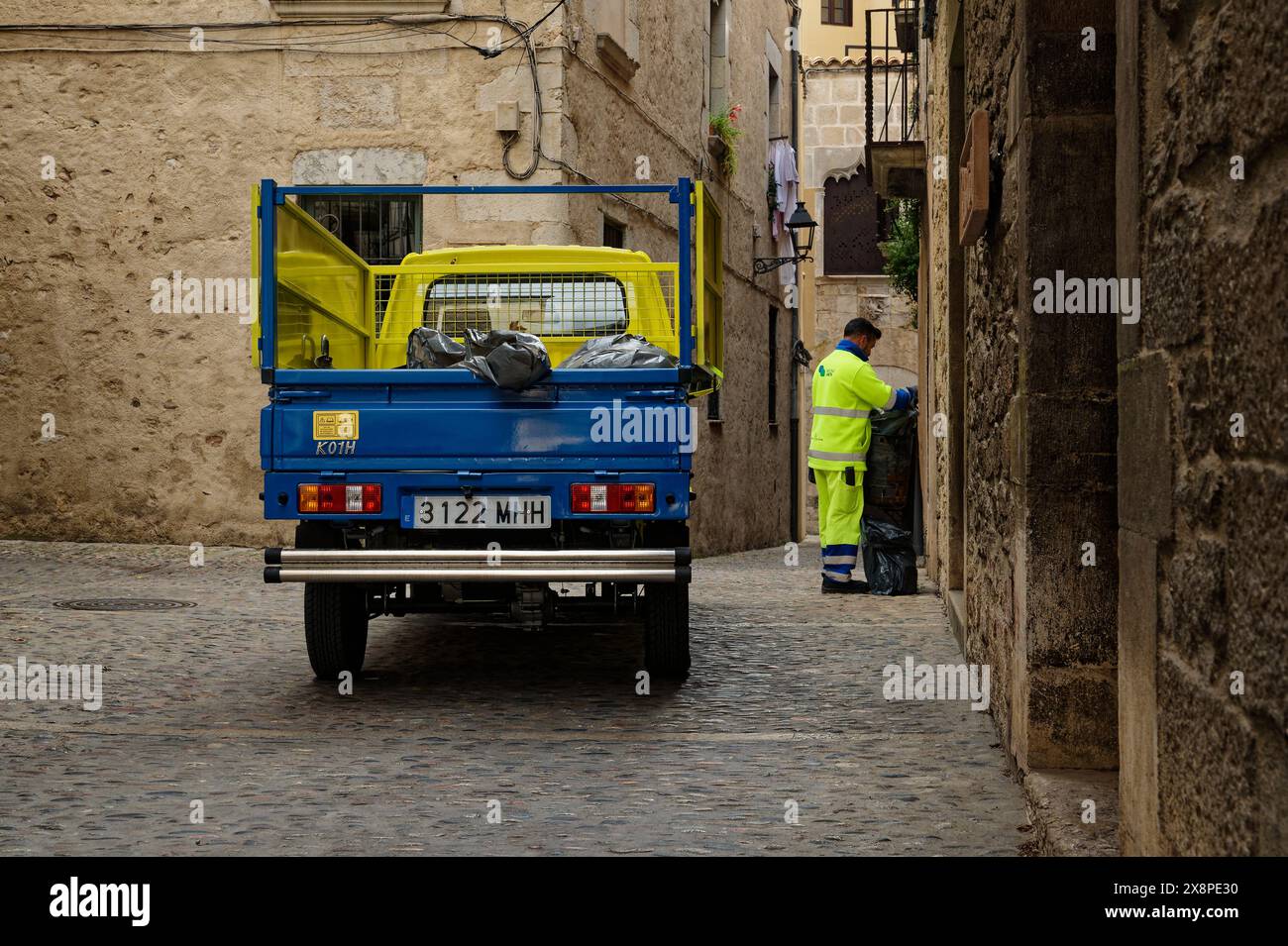 Un lavoratore municipale che raccoglie la spazzatura da una stretta strada acciottolata della città di Girona, con un camion colorato di colore blu e giallo parcheggiato. Foto Stock