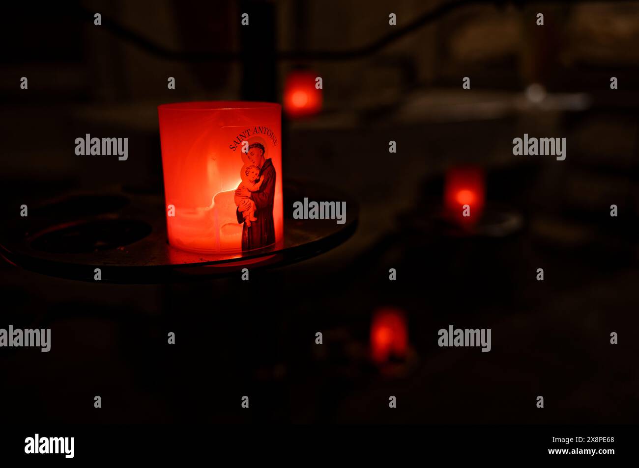 In una chiesa poco illuminata a Carcassonne, una candela rossa raffigurante un santo sfarfallio, con altre candele soffuse sullo sfondo. Foto Stock