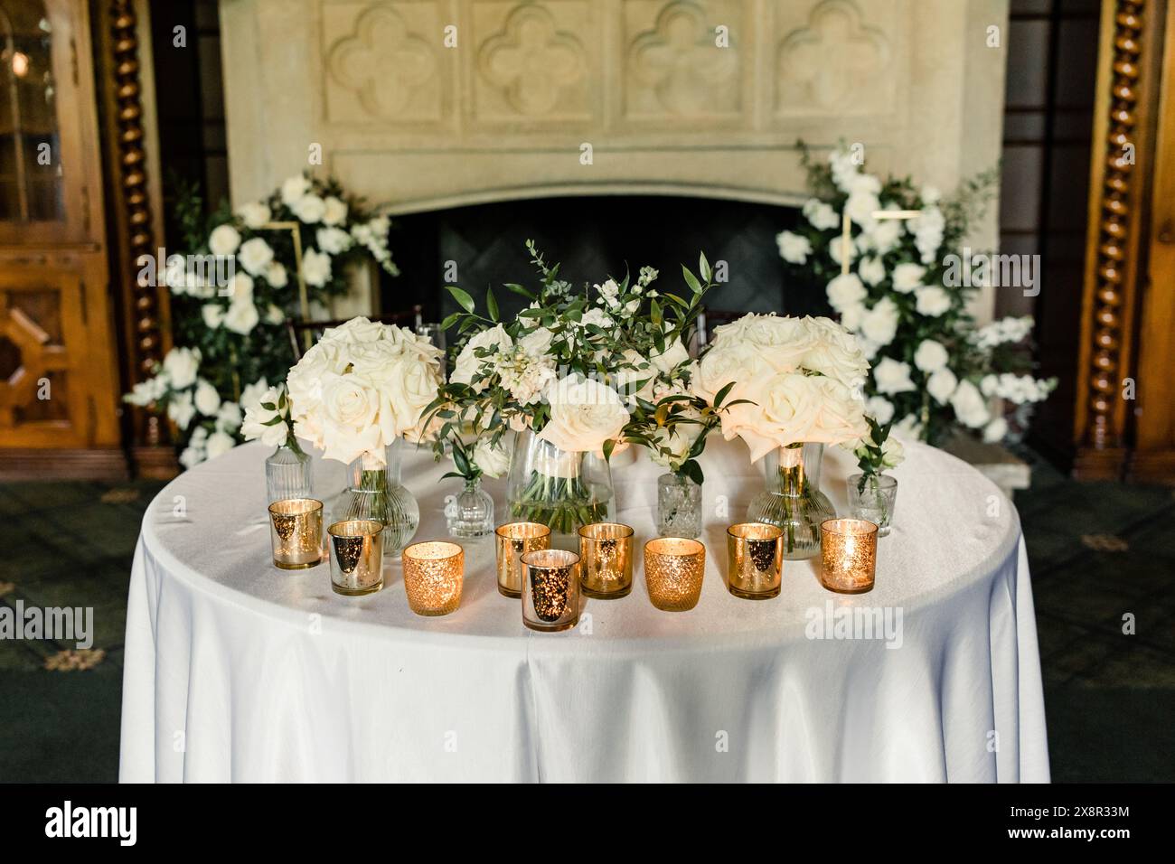 Tavolo con rose bianche e candele votive dorate davanti a un caminetto Foto Stock
