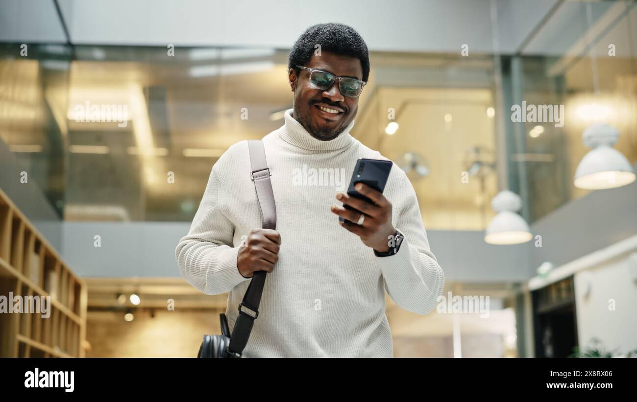 Ritratto del giovane Black Man che si sente sicuro di sé mentre cammina sorride e controlla il suo telefono, in un corridoio occupato dell'ufficio. Sta navigando in Internet sul suo smartphone. Angolo basso Foto Stock