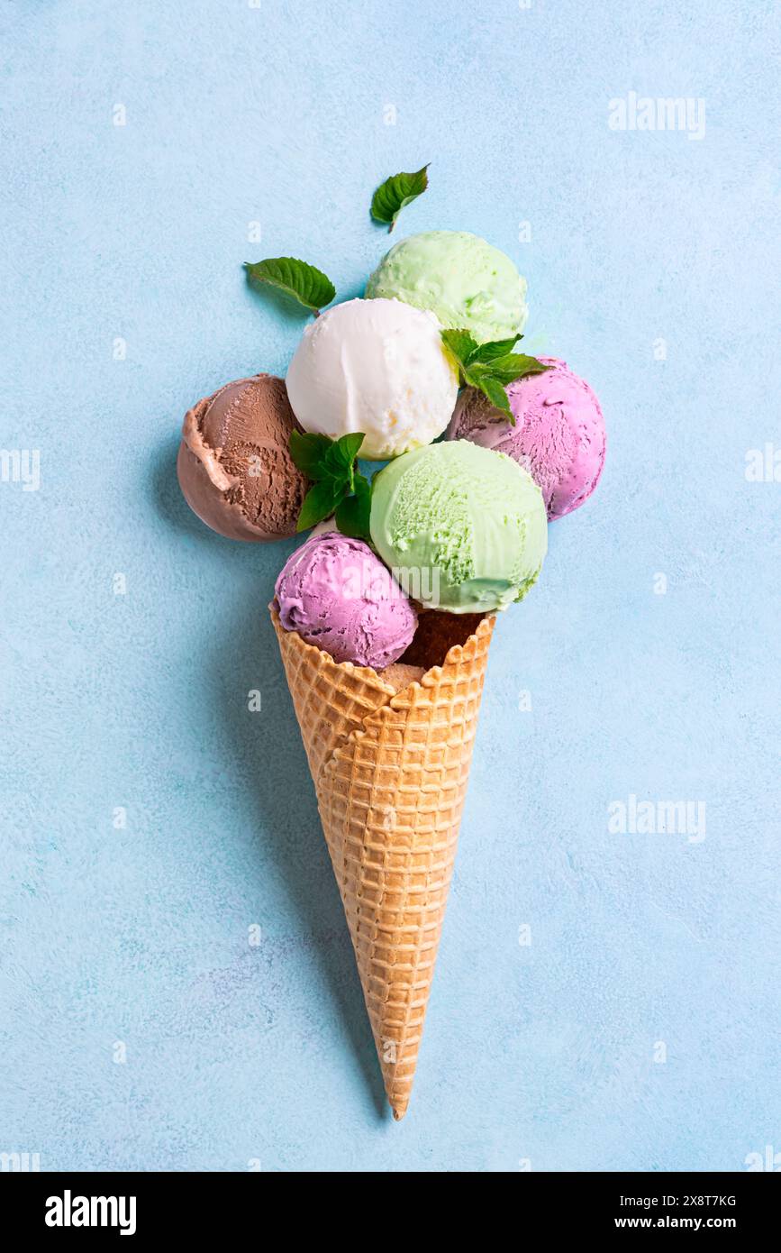 Cono per gelato con vari cucchiai su sfondo blu, piatto e creativo. Molti gelati biologici colorati nel cono per waffle. Foto Stock
