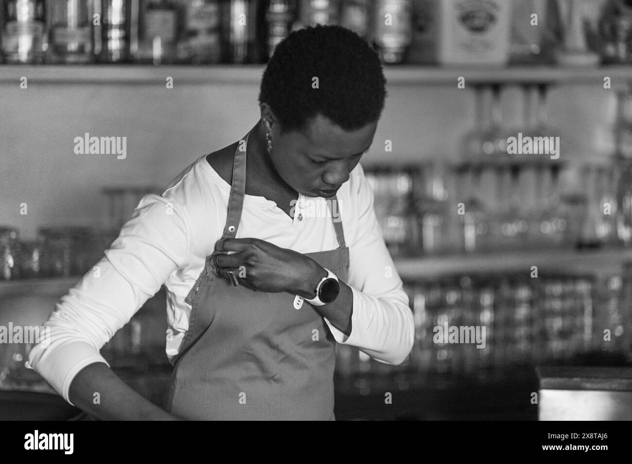 Il barista concentrato che indossa un grembiule si trova di fronte a una varietà di bottiglie in un bar pieno Foto Stock