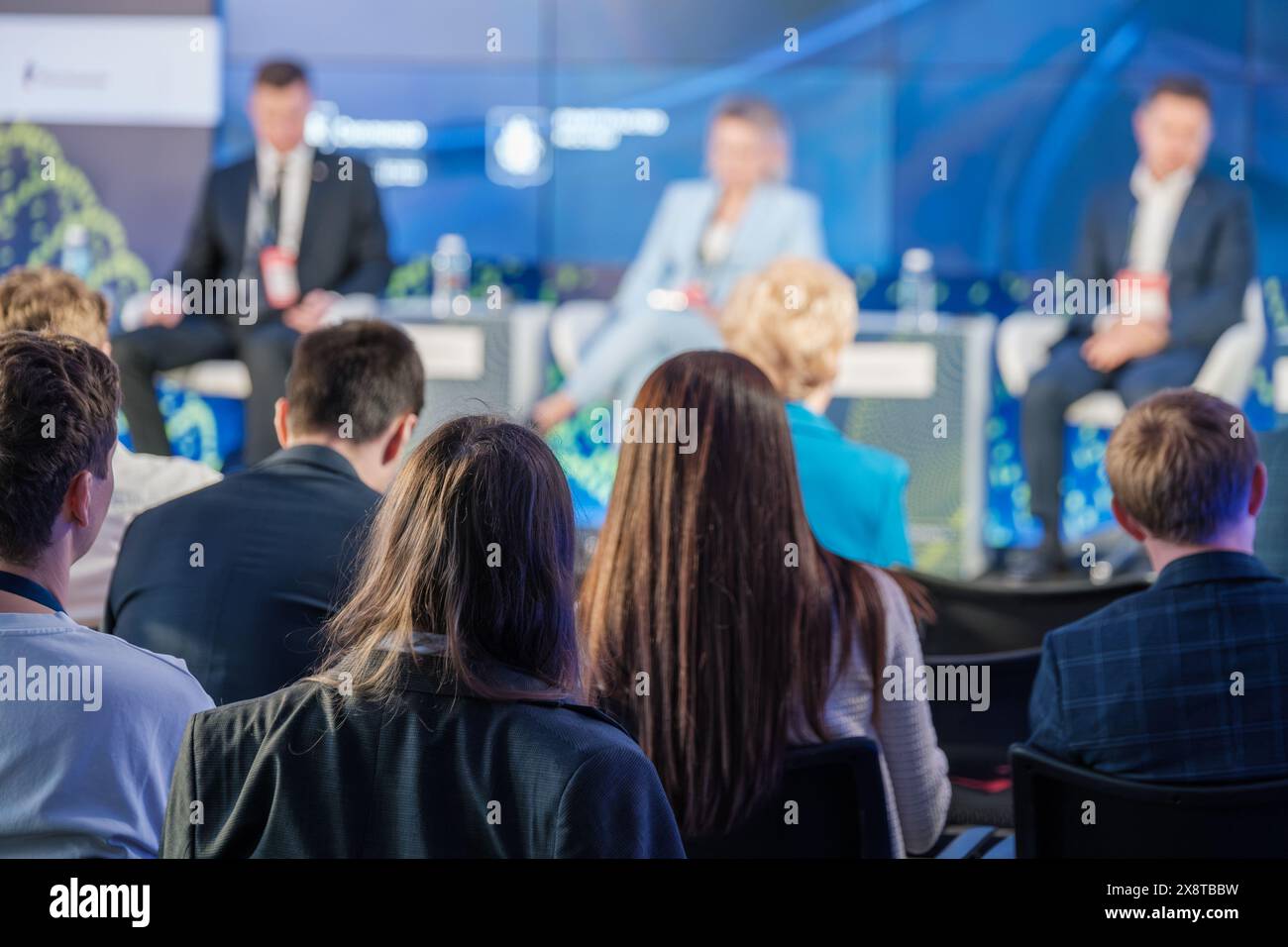 Un pubblico diversificato ascolta attentamente gli oratori sul palco durante una conferenza di lavoro. I panelisti appaiono sfocati mentre discutono gli argomenti chiave. Foto Stock