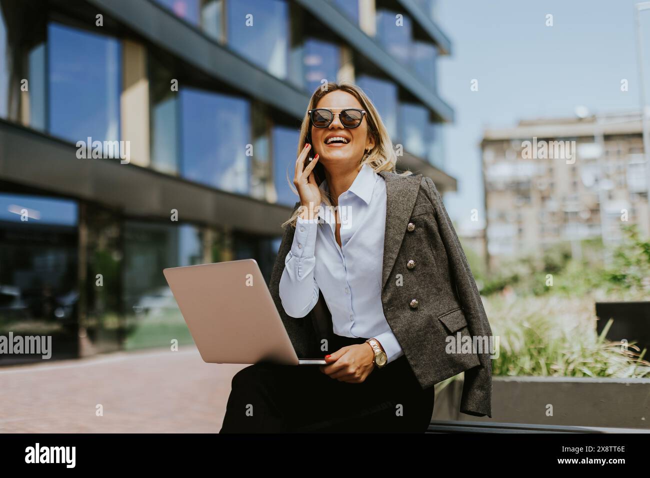 Un professionista sorridente che usa il suo smartphone e il suo laptop fuori da un elegante edificio moderno per uffici in un pomeriggio di sole. Foto Stock