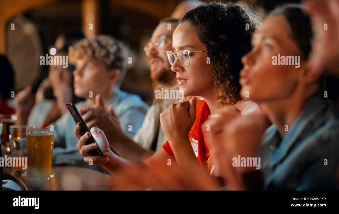 Ritratto di un'ansiosa donna multietnica nella maglia rossa con uno smartphone, nervosa per la scommessa sportiva sulla sua squadra di calcio preferita. Giovane donna piena di gioia quando la squadra di calcio segna un gol. Foto Stock